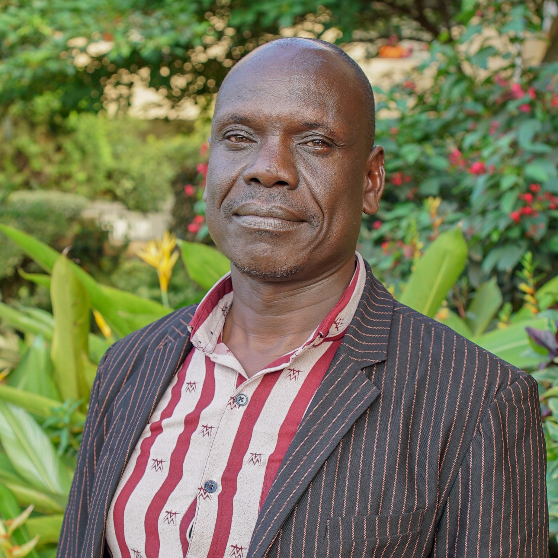 James Peter Inyangat James Peter Inyangat in a dark pinstripe blazer over a red striped shirt, standing in a lush garden with tropical plants and colorful blooms in the background.