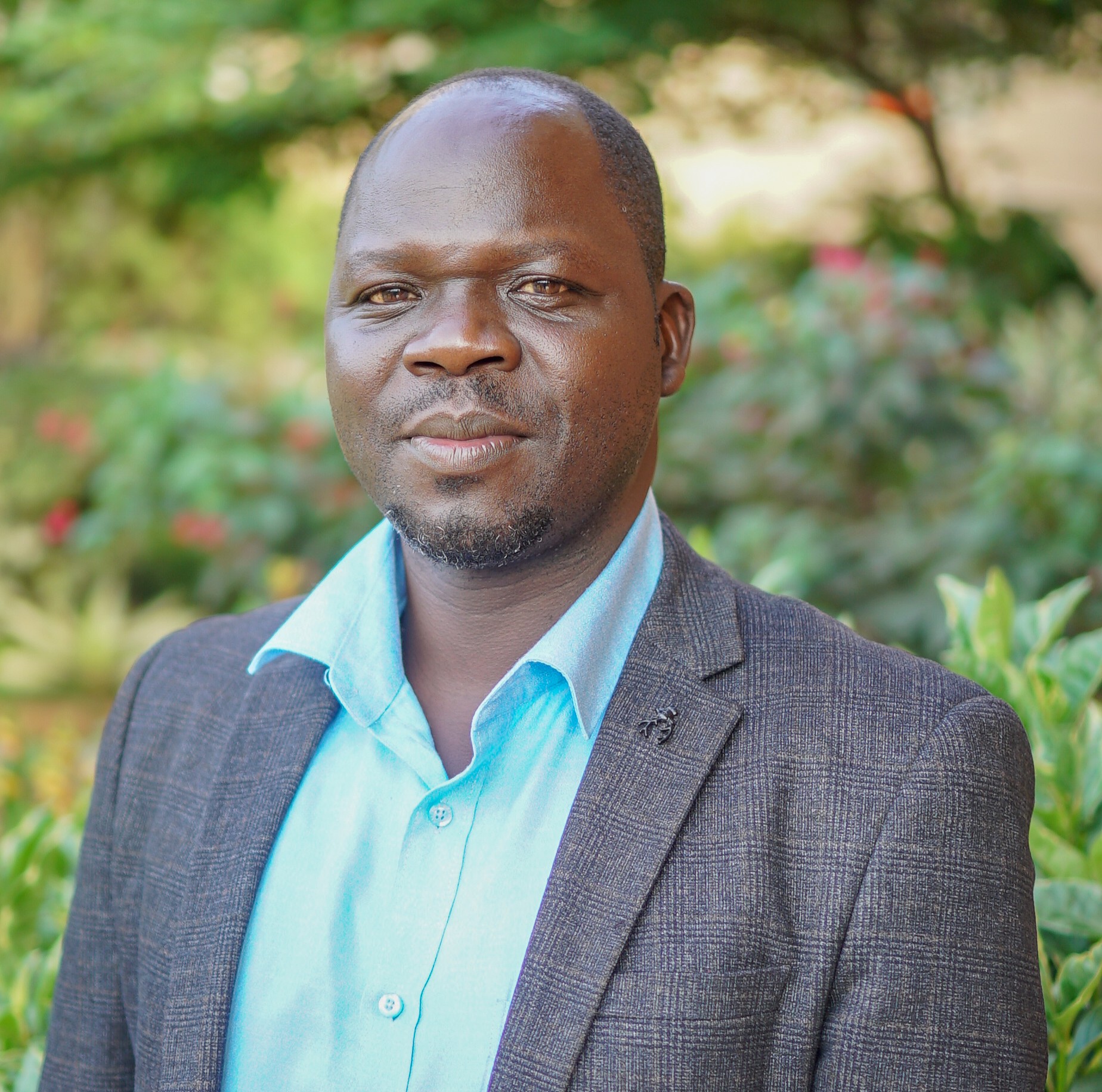 Kenneth Odur Kenneth-Odur wearing a light blue shirt and a dark textured blazer outdoors, with lush green foliage and soft-focus flowers in the background