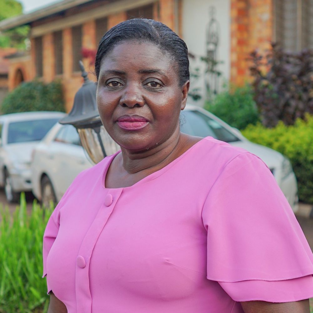 Najjuma Irene wearing a pink short-sleeve blouse, photographed outdoors with greenery and a car in the background.