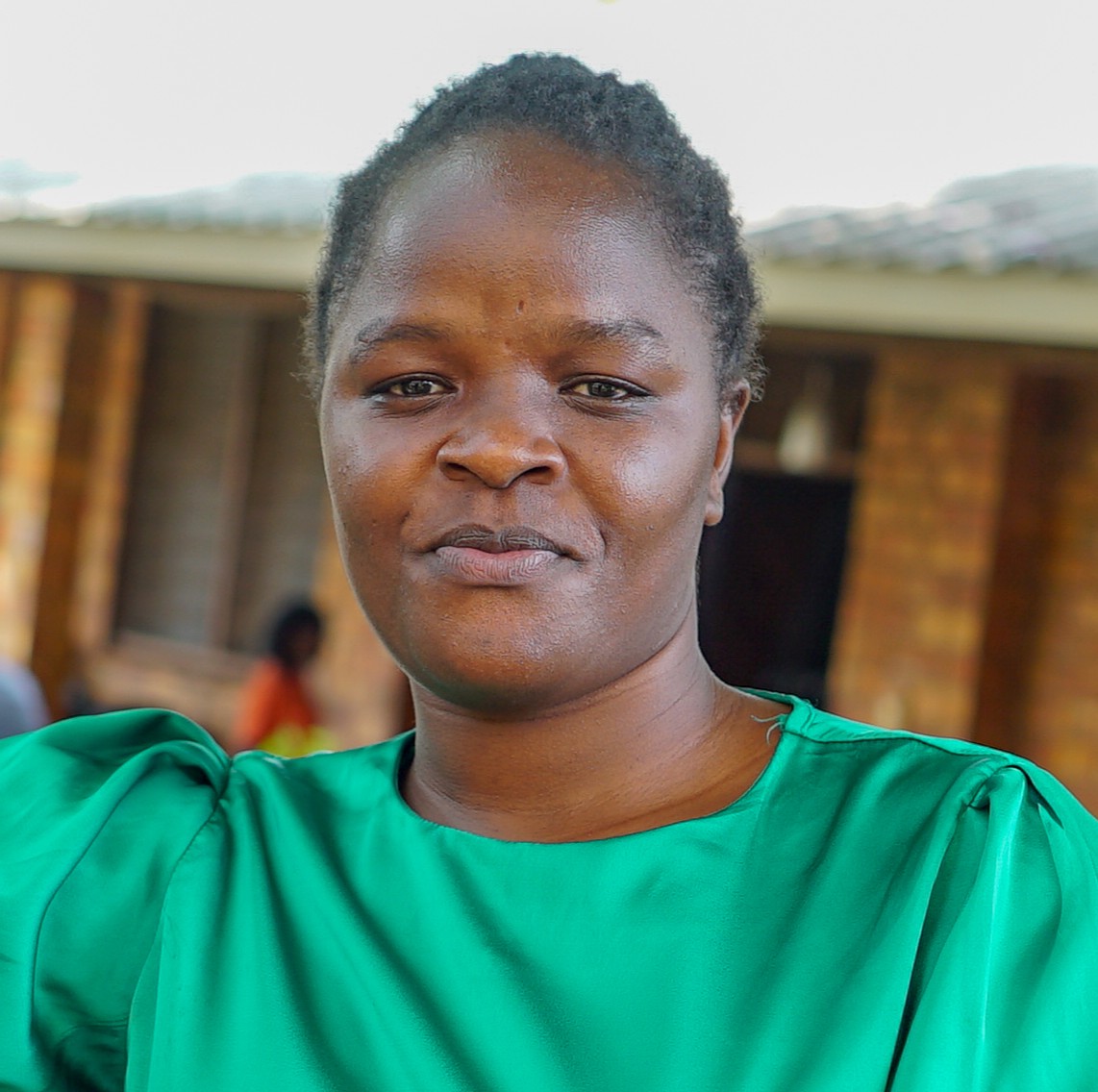 Namono Jackline Jackline in a bright green dress stands outdoors with a calm expression, short natural hair, brown skin, and a brick building backdrop.