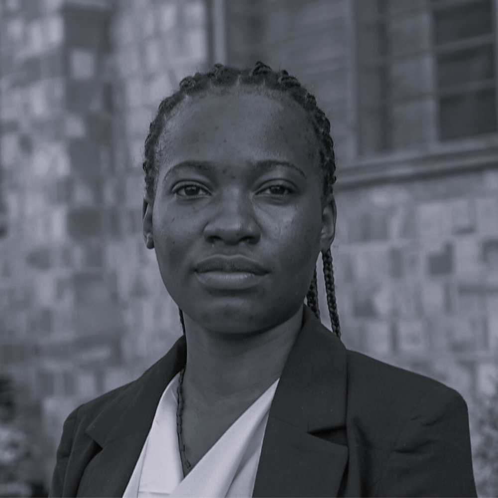 Ndagire Angella, Administrative Officer, in a dark blazer with a white blouse, portrait-style against a brick wall backdrop, Ugandan professional.