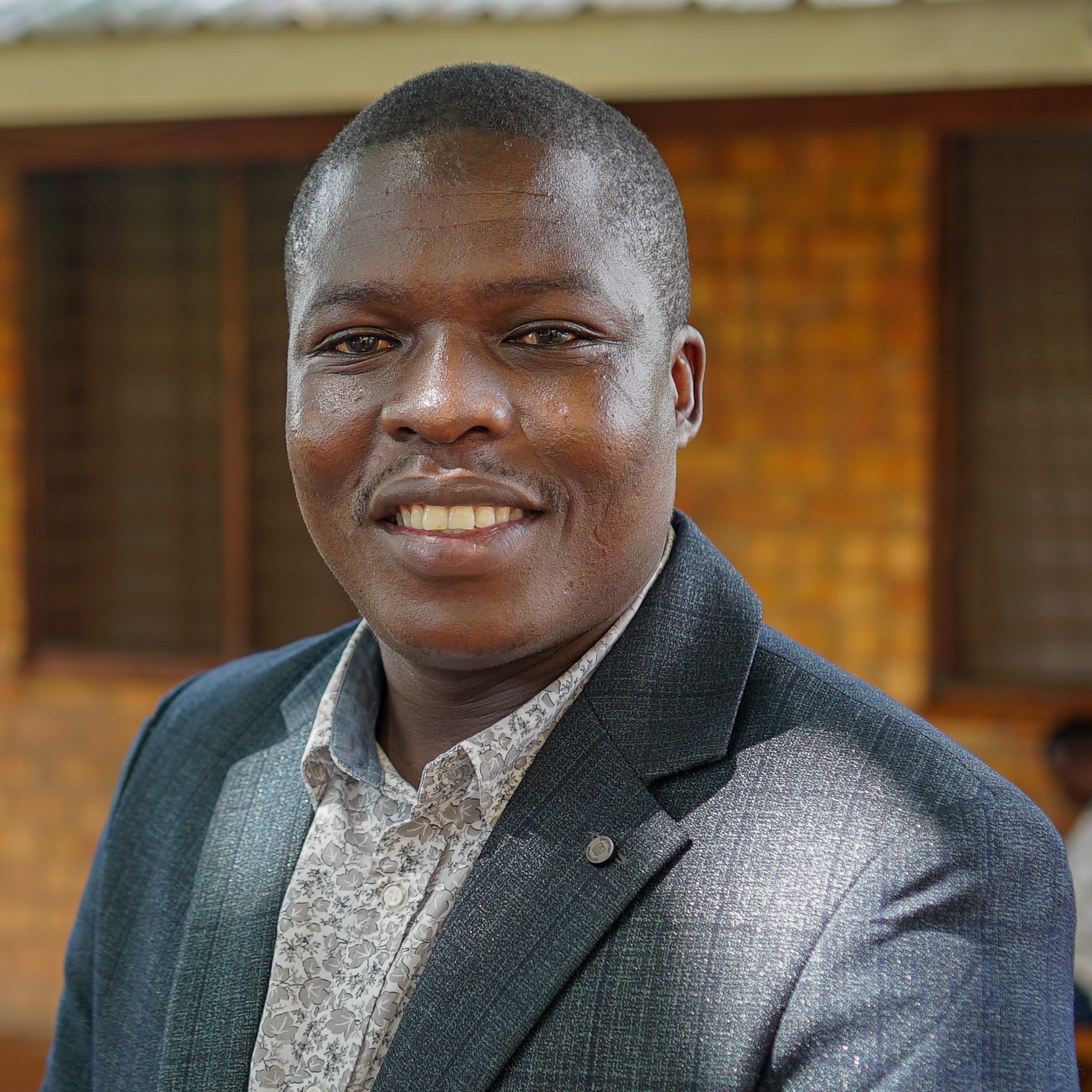 Omondi Eric Omondi-Eric smiling man in a dark gray blazer and light patterned shirt, standing outdoors with a warm brick wall backdrop and soft daylight.