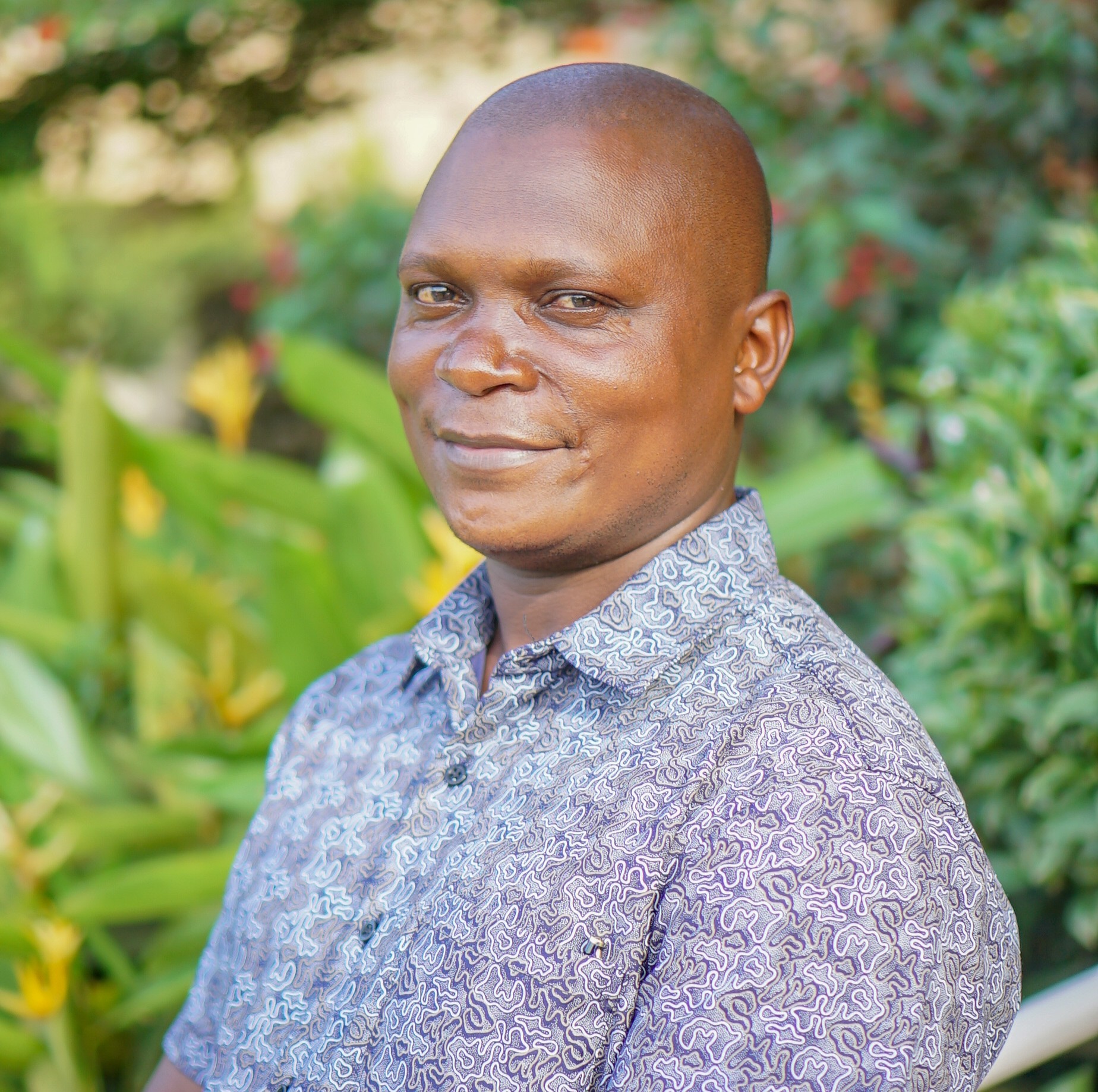 Wangira_Denis Wangira Denis stands outdoors in a lush garden wearing a blue-grey patterned short-sleeve shirt and a warm smile, with a shaved head and soft sunlight on his face.