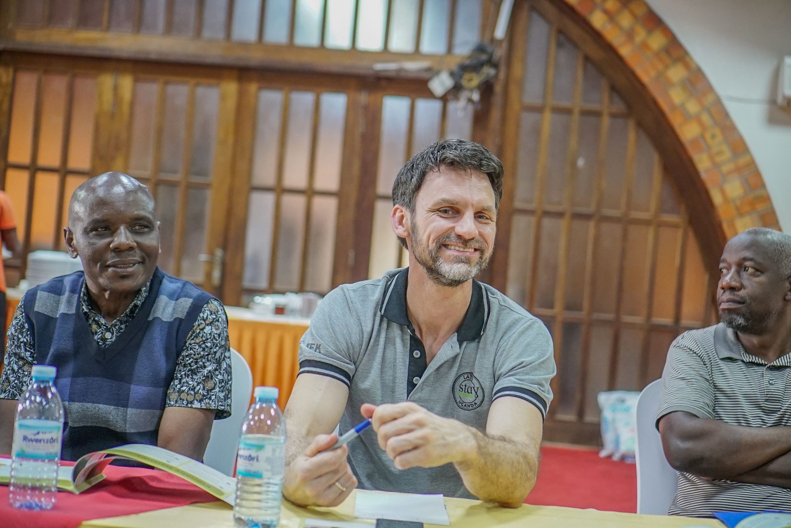 Men seated at a meeting table with water bottles, smiling in front of a wooden hall backdrop and a gray polo shirt with Stay Alliance logo.