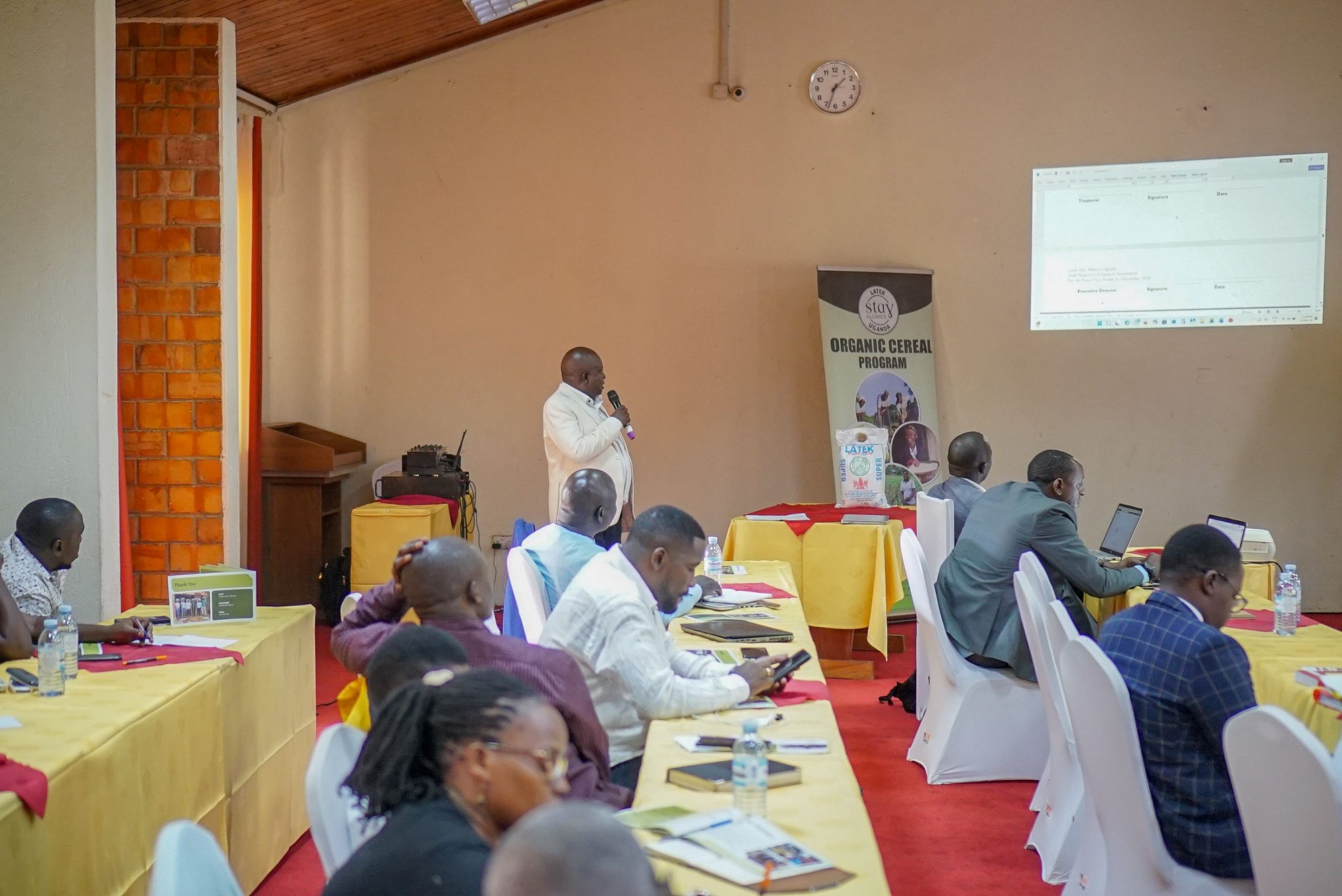 A community training session with a speaker holding a microphone, yellow tablecloths, an “ORGANIC CEREAL PROGRAM” banner, and laptops in a brick-walled room.