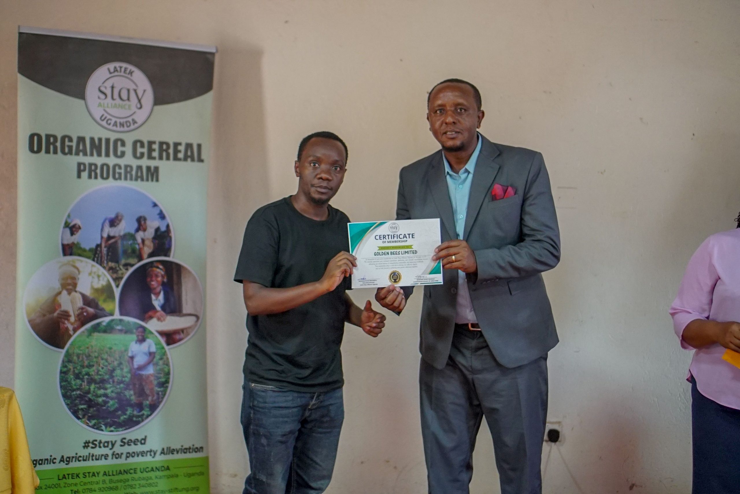 A Stay Alliance Uganda certificate presentation beside a poster for the Organic Cereal Program, with Golden Bees Limited named on the document