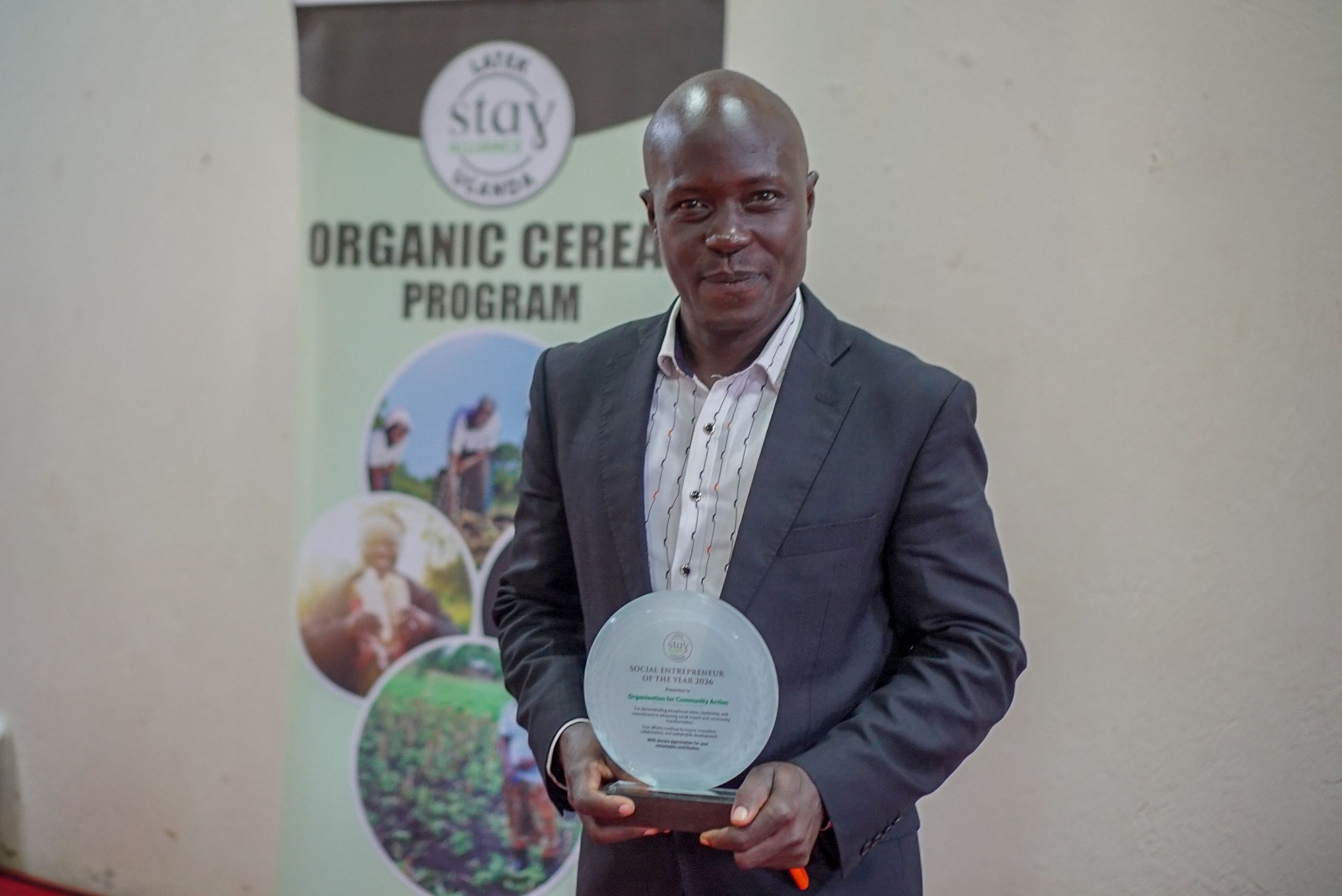 Man in a black suit and white shirt holds a Stay Alliance award plate in front of a backdrop reading Organic Cereal Program.