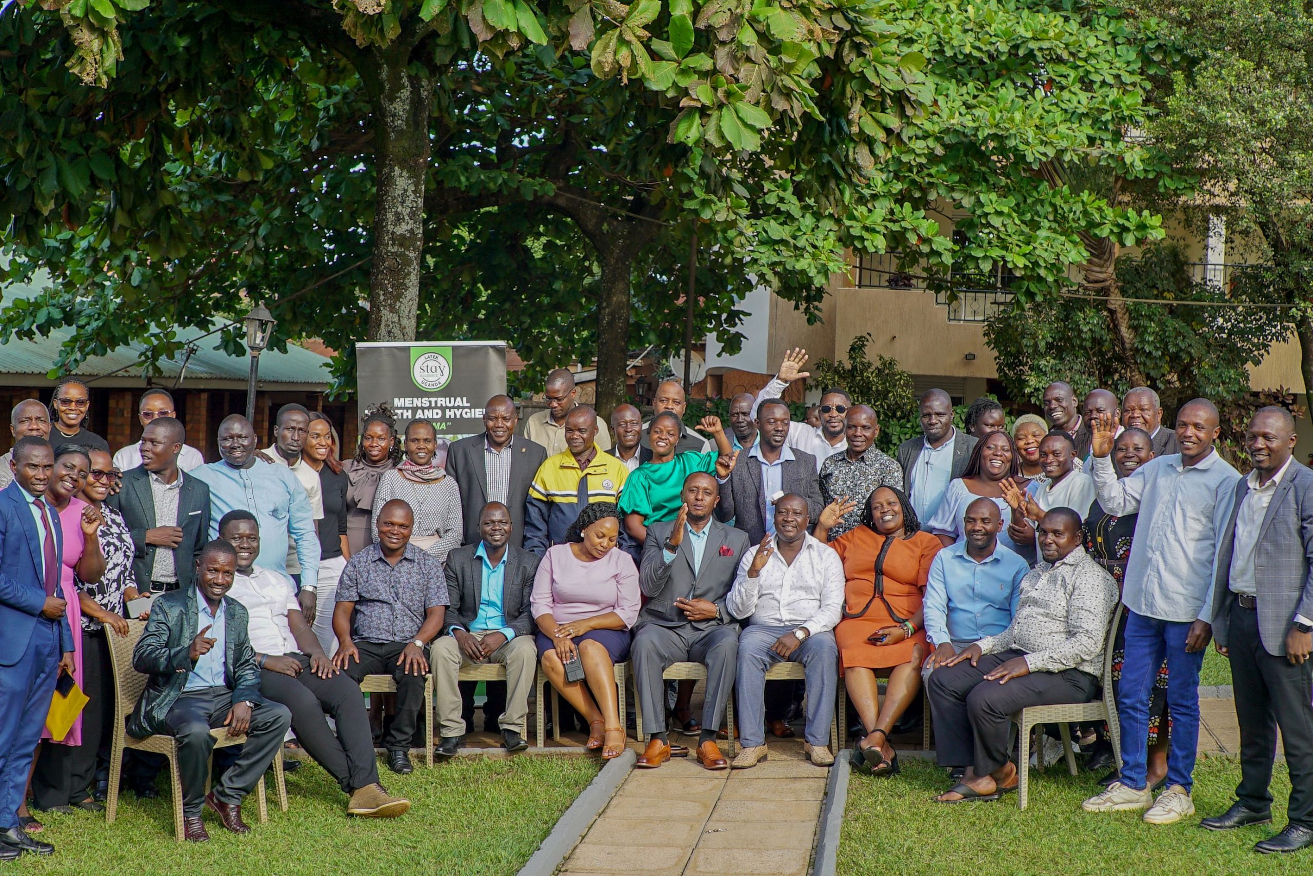 Group photo outdoors with green foliage overhead and a banner reading “Menstrual Health and Hygiene” from Stay Alliance.