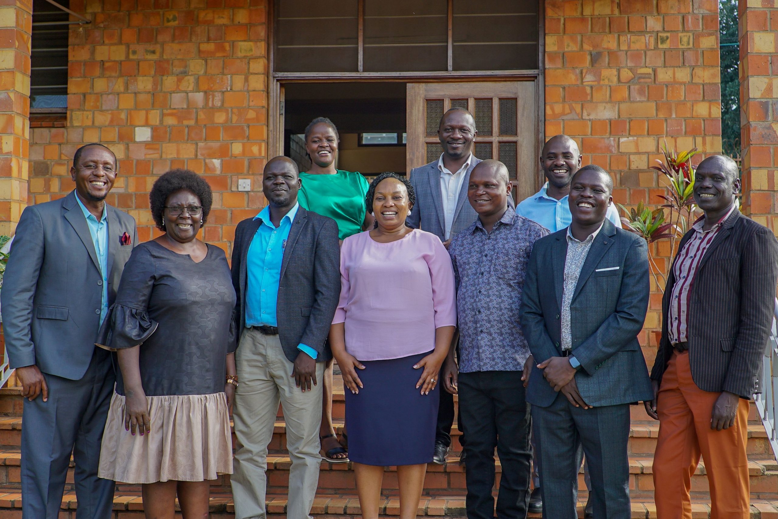 Smiling team members in suits and dresses pose in front of a red-brown brick building entrance, including one person in a green top.